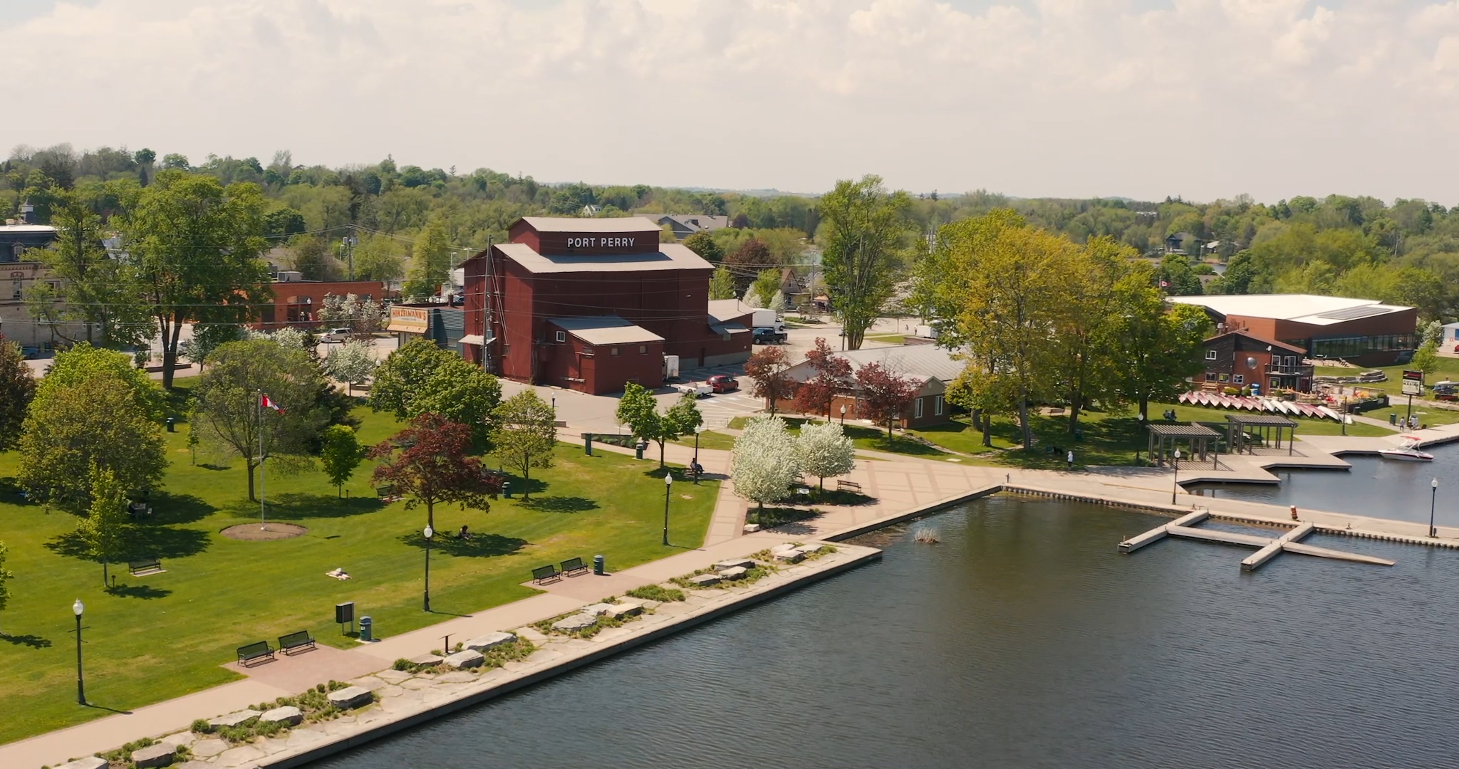 A large aerial shot of Lake Scugog in Downtown Perry. Palmer Park is a large greenspace along the waterfront featuring picnic areas, playgrounds and benches. The marina and Old Mill can be seen in the background. Streets connect to the Historic downtown of Port Perry.