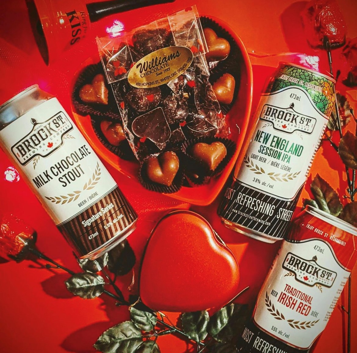 Three Brock St. Brewing beer cans on a red background with Williams Chocolate hearts, roses and a large red heart