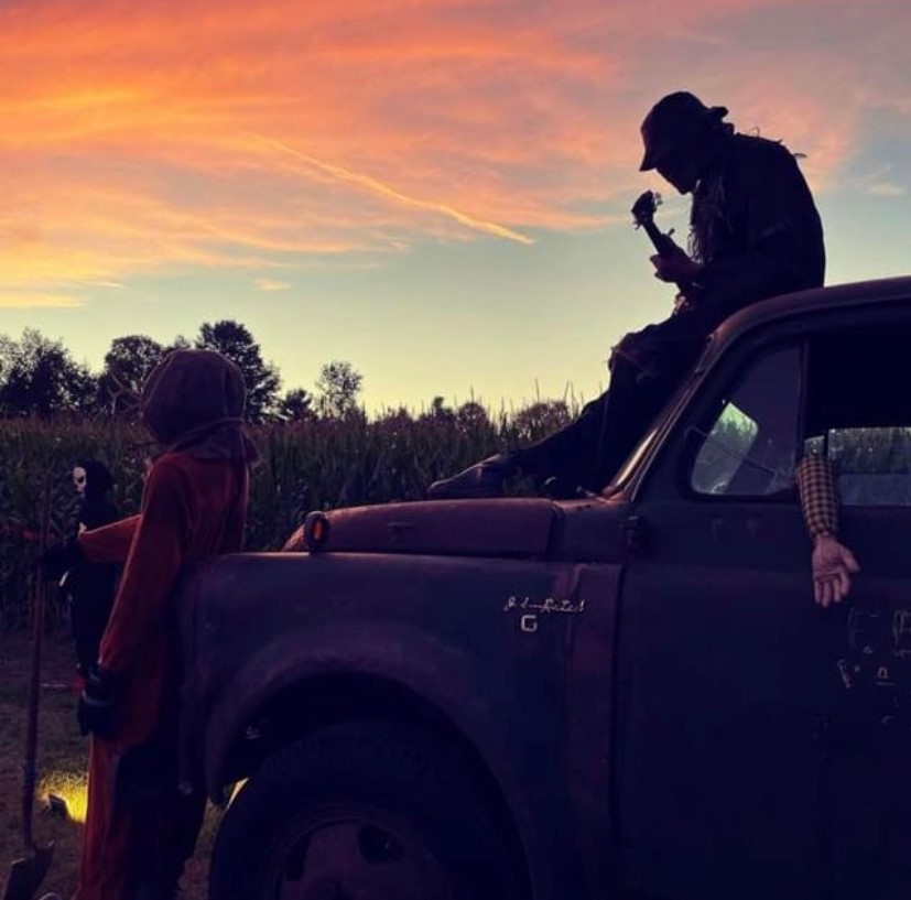 Image of a corn field under an orange sky with props including skeletons and an old truck.