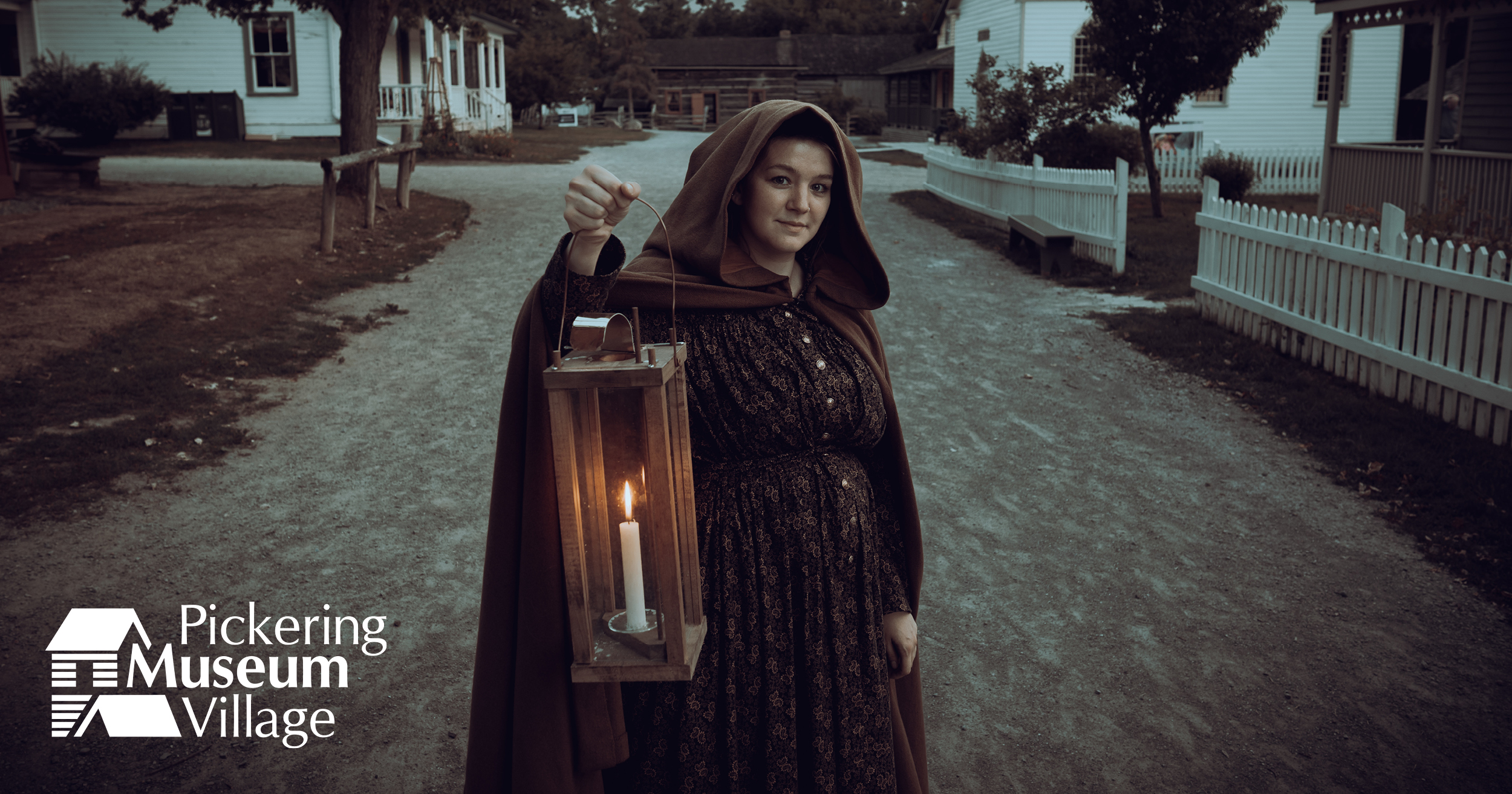 Image of a woman wearing a black cloak and holding a lantern in front of old buildings.
