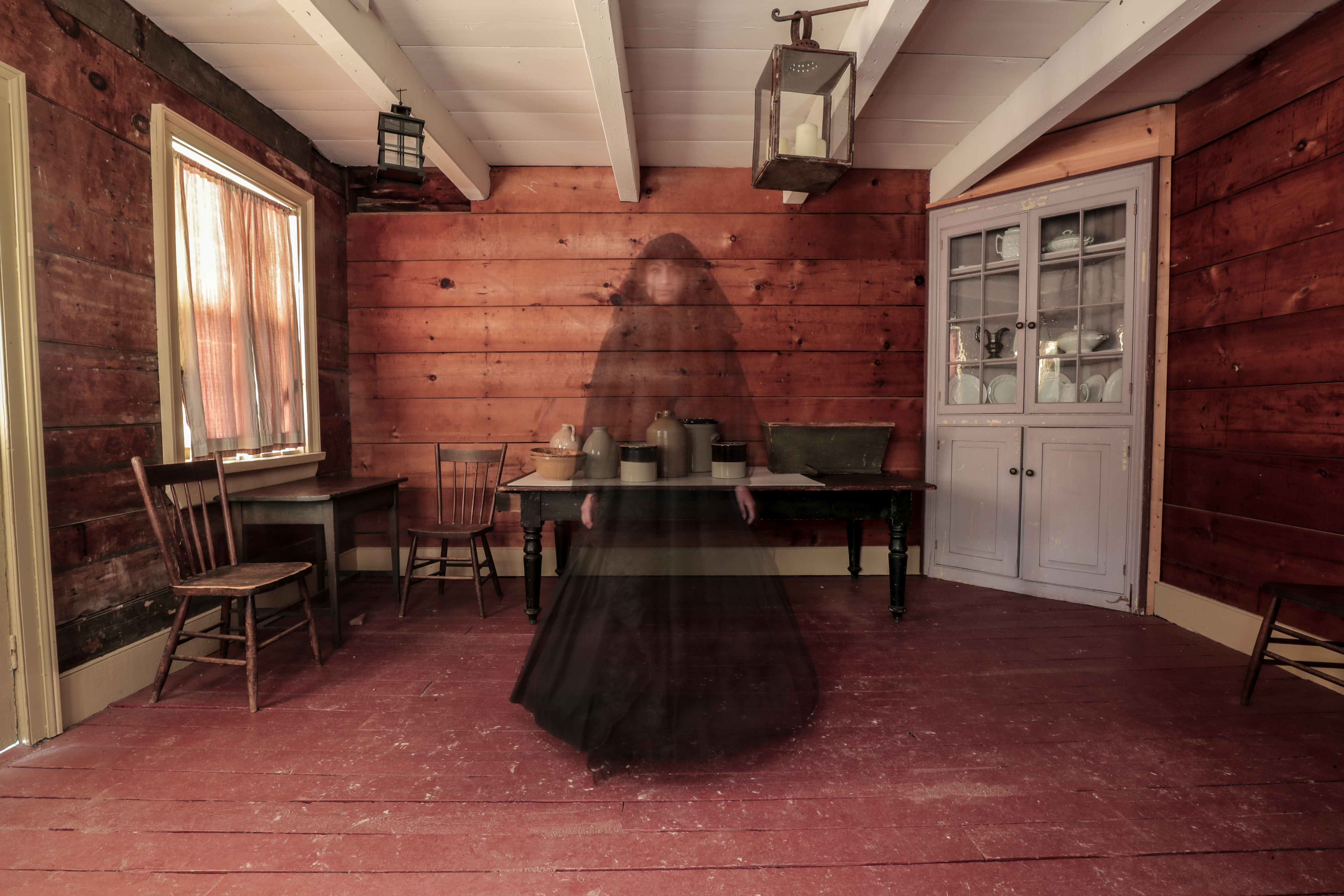 Image of a shadowy figure in a black dress standing inside an old log cabin.