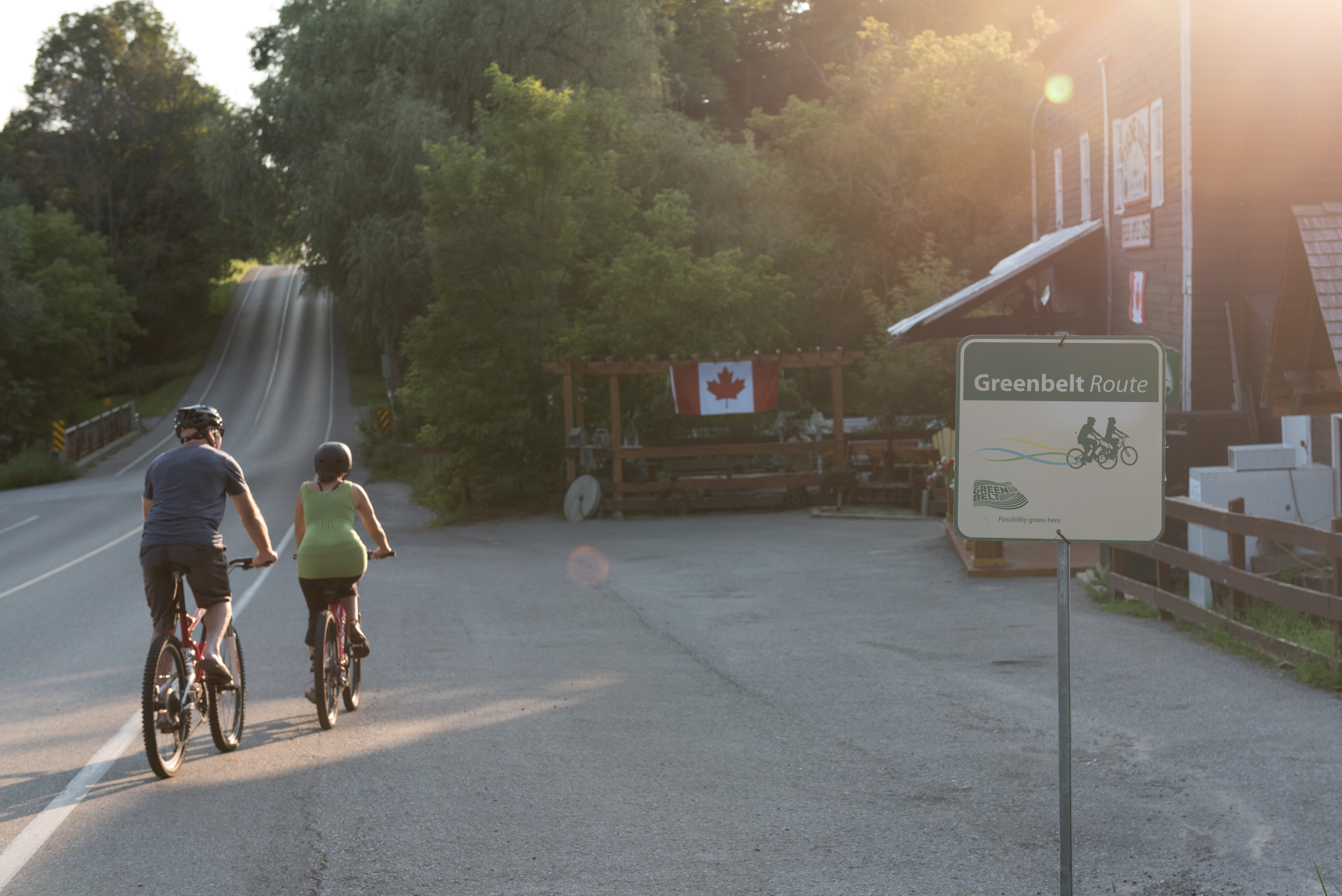 Two cyclists riding their bikes and arriving at Tyrone Mills in Clarington. There is a Greenbelt Cycling route sign, 