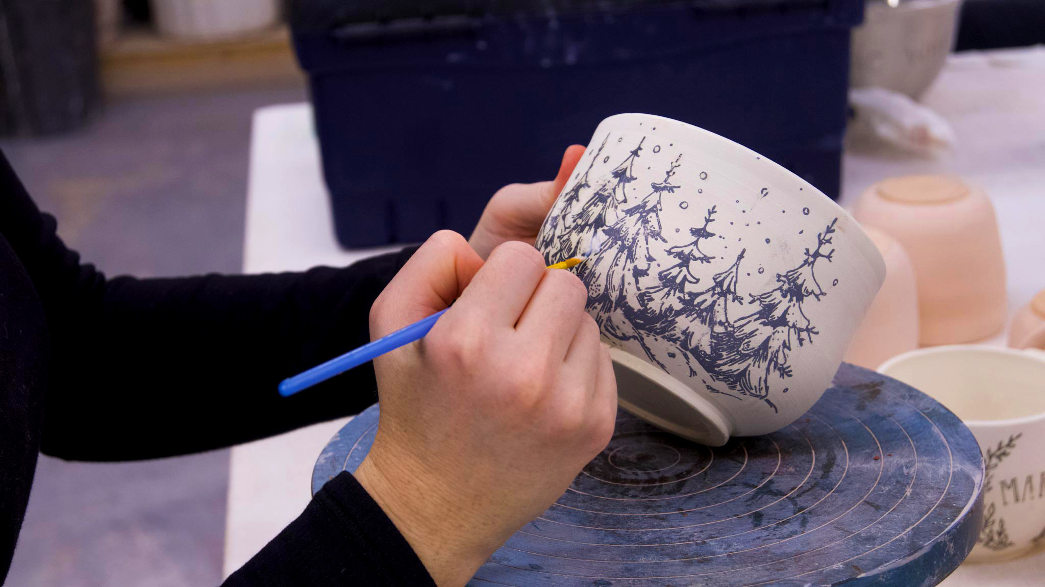 Hands painting a ceramic cup with blue trees