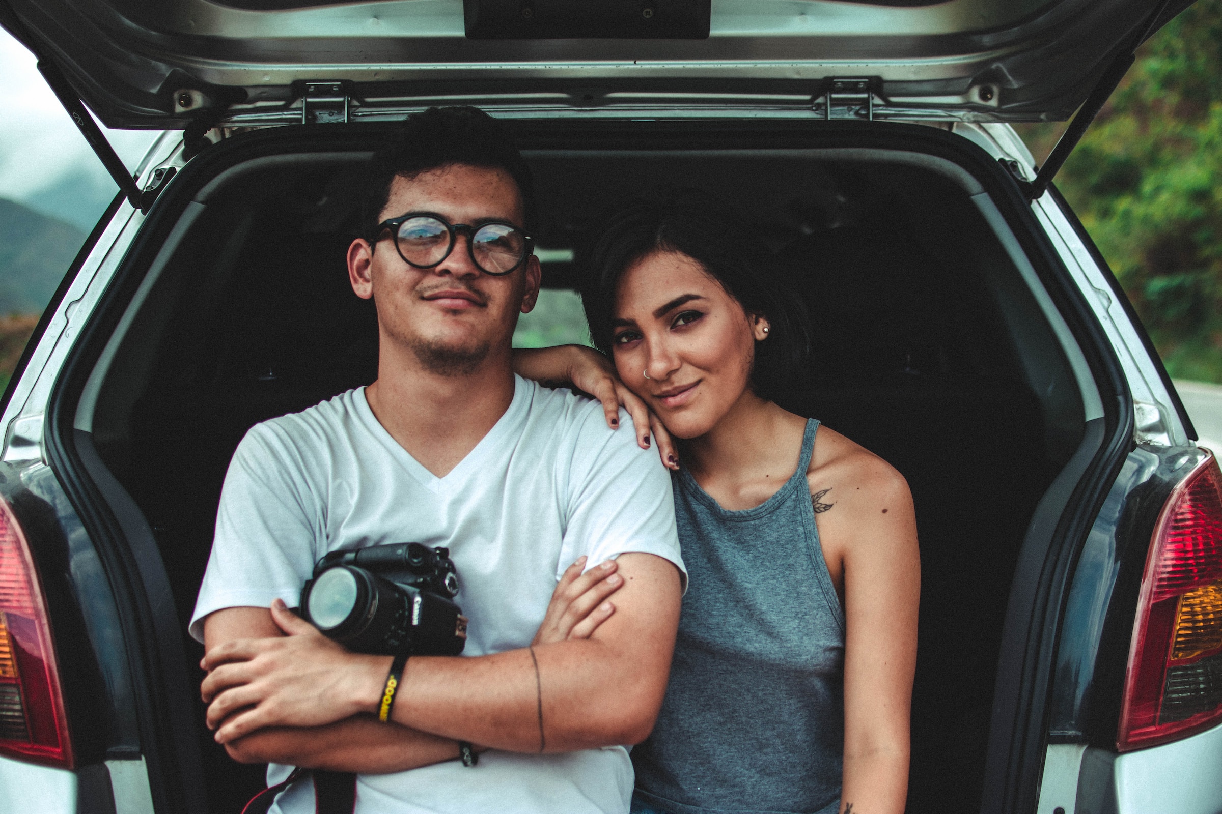 A couple leaning on the back of their vehicle holding a camera