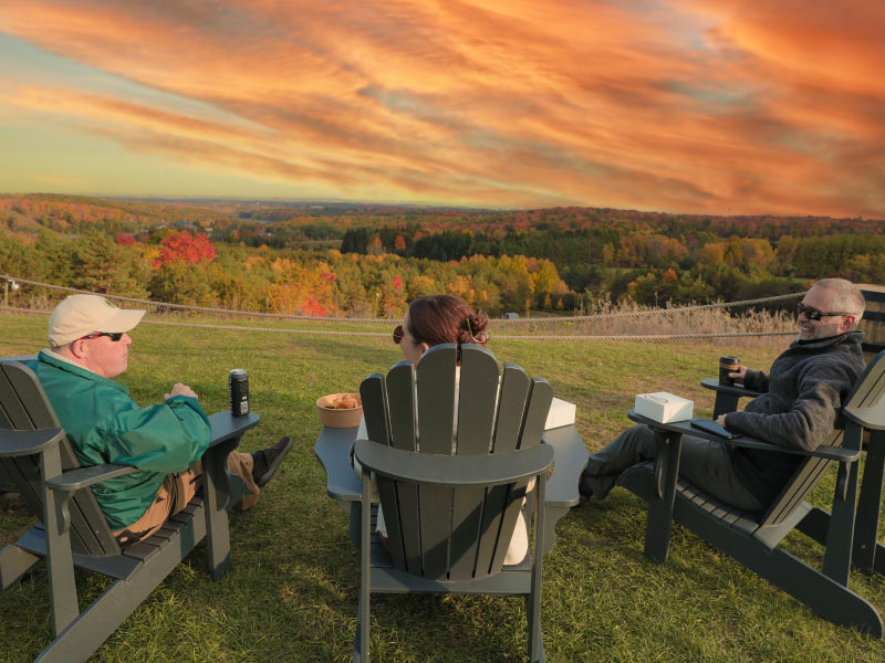 Three people sitting in Adirondack chairs overlooking the fall trees and a sunset.