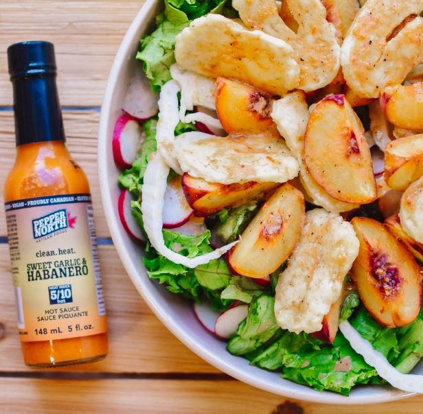 A close up of a small bottle of Pepper North hot sauce beside a salad with peaches.