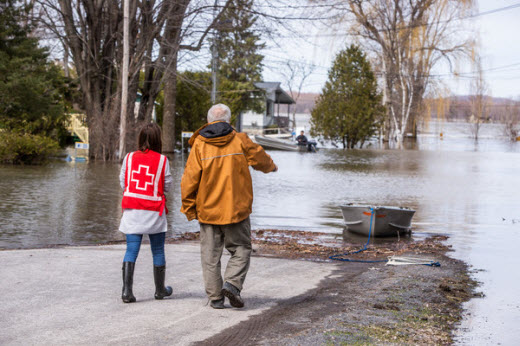 Two people walking towards a flooded road, one wearing a red vest with the red cross symbol