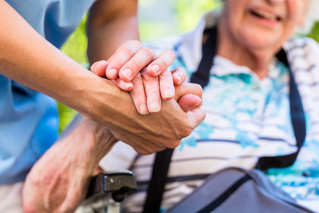 Two people holding hands with a seated elderly person being supported by a standing person in the background