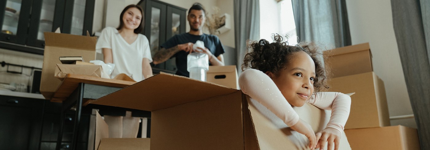 child sitting in a box as parents look on from the kitchen