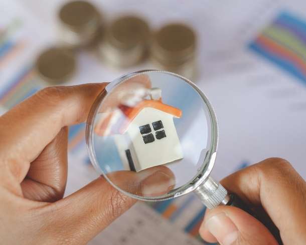 Hands holding magnifying glass over miniature house. Coins and graphs out of focus in the background.