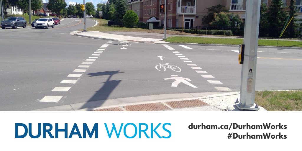 : Image shows a crossride made of white dashes painted on roadway spanning across the road to connect two multi-use paths. An icon of a person walking and a bike are also painted on the road. Text under image states: “Durham Works, durham.ca/DurhamWorks #DurhamWorks.”