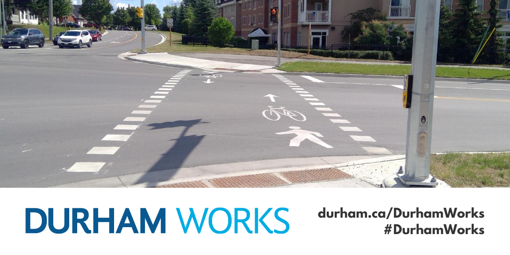 Image shows a crossride made of white dashes painted on roadway spanning across the road to connect two multi-use paths. An icon of a person walking and a bike are also painted on the road. Text under image states: “Durham Works, durham.ca/DurhamWorks #DurhamWorks.