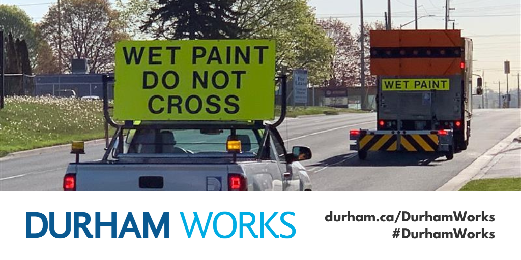 A construction truck with a construction arrow indicating that drivers need to go around it, followed by a pickup truck with a sign that says, Wet paint. Do not cross. Text below image states, “Durham Works, durham.ca/DurhamWorks, #DurhamWorks.”