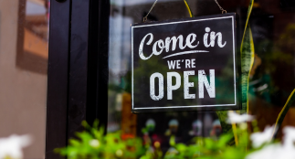 An image of a store window with a sign hanging and white text that reads “Come in we’re open”.