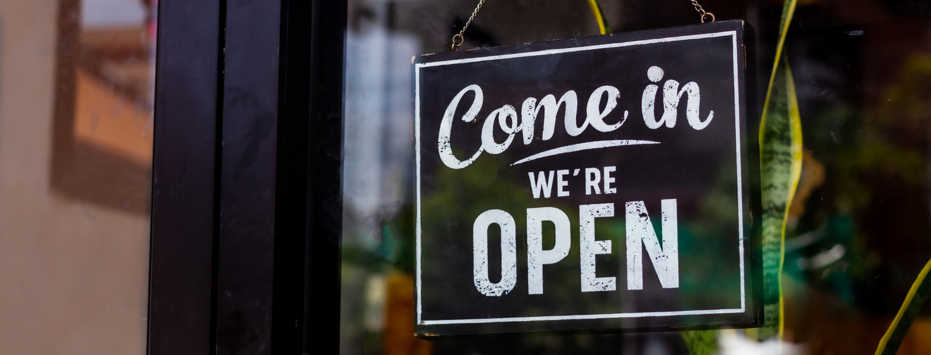 An image of a store window with a sign hanging and white text that reads “Come in we’re open”.