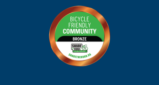A circular “Bicycle Friendly Community – Bronze” badge with a green and white center and a bronze outer ring. The badge includes the Share the Road logo and the website ShareTheRoad.ca, displayed on a dark blue background.