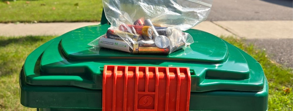 A clear bag with used batteries in it on top of a green bin. 