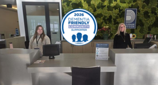 A customer service counter at a Service Durham location with two staff members standing behind separate workstations. A large circular badge displaying “2026 Dementia Friendly Community Supporter” is centered above the counter. The background features a green living wall design and a Service Durham sign on the right.