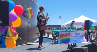 A person on a stage speaking into a microphone with Pride Balloons in the background.