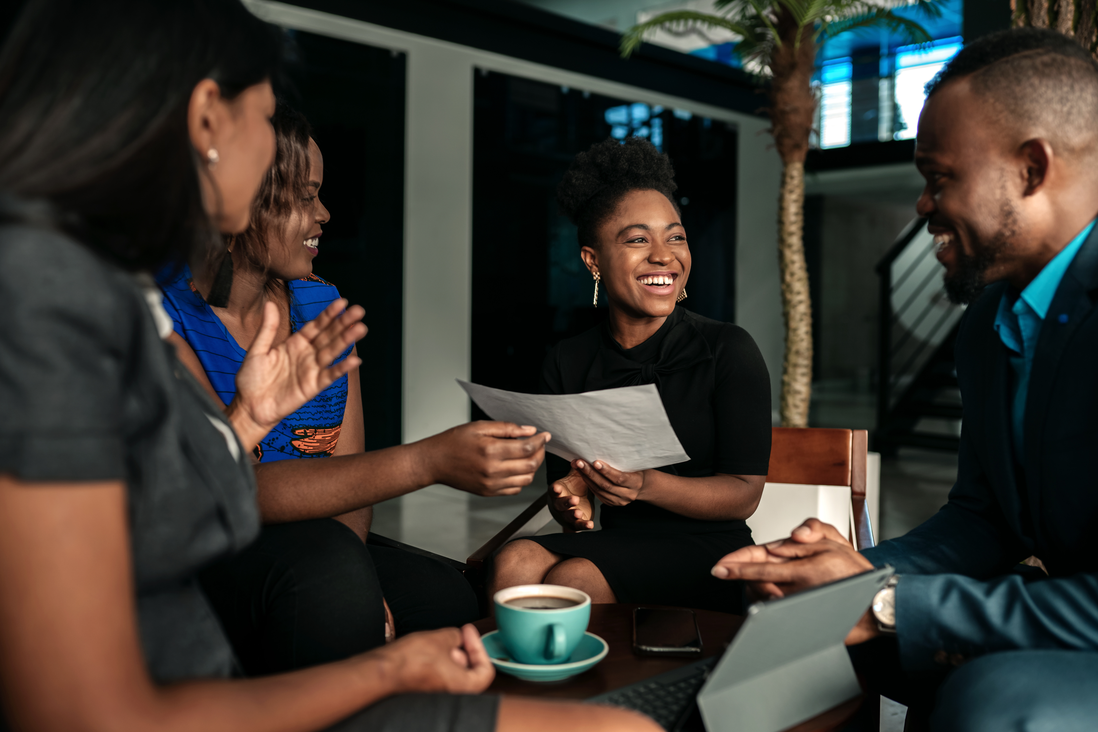 A group of people sitting in a circle, looking at each other and smiling. One person holds a piece of paper, another has a drink sitting on their lap.