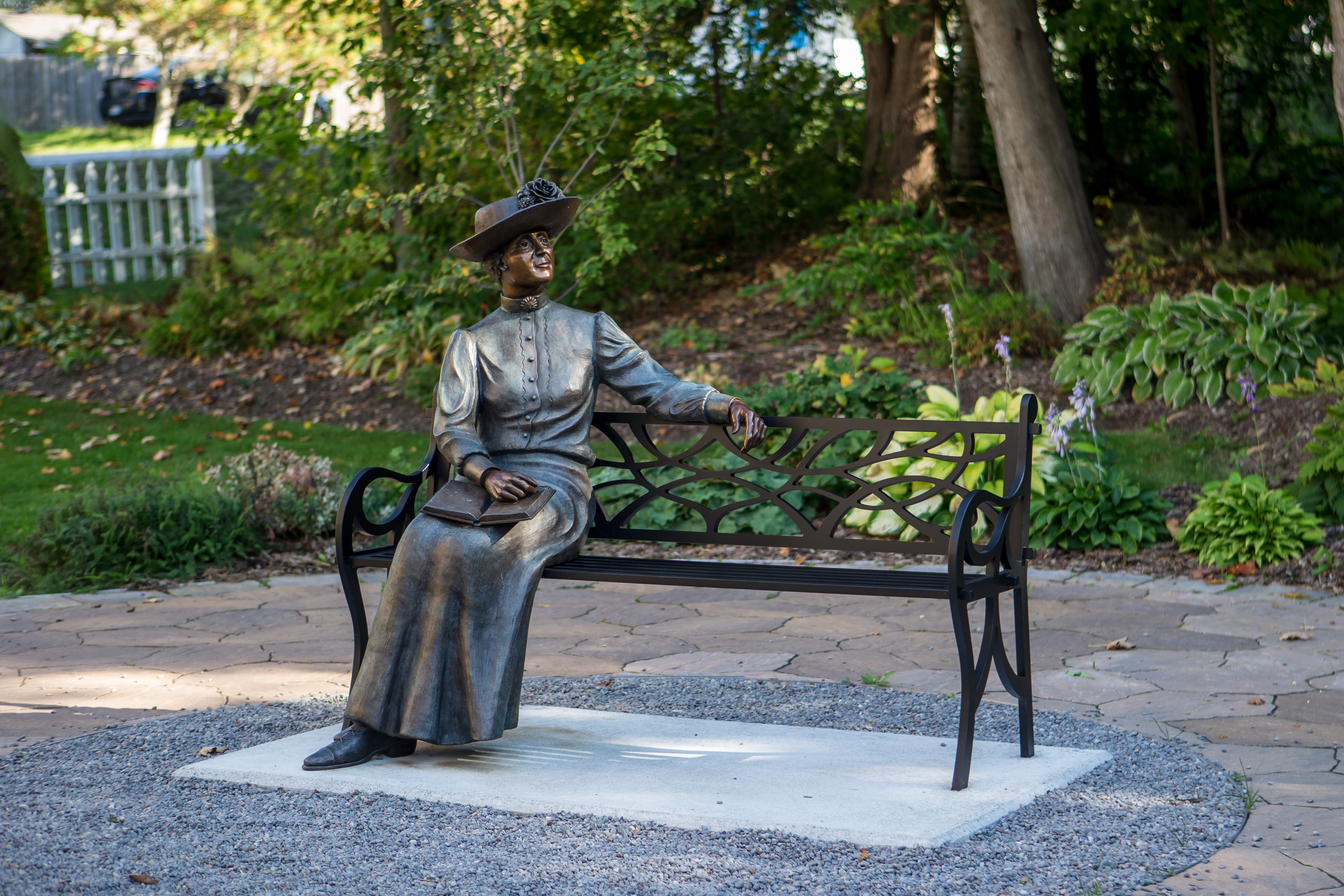 Image is of a bench that has a statue of Lucy Maud Montgomery with a book on her lap sitting on the left side of the bench. The right side of the bench is empty and is located in front of a garden and trees.