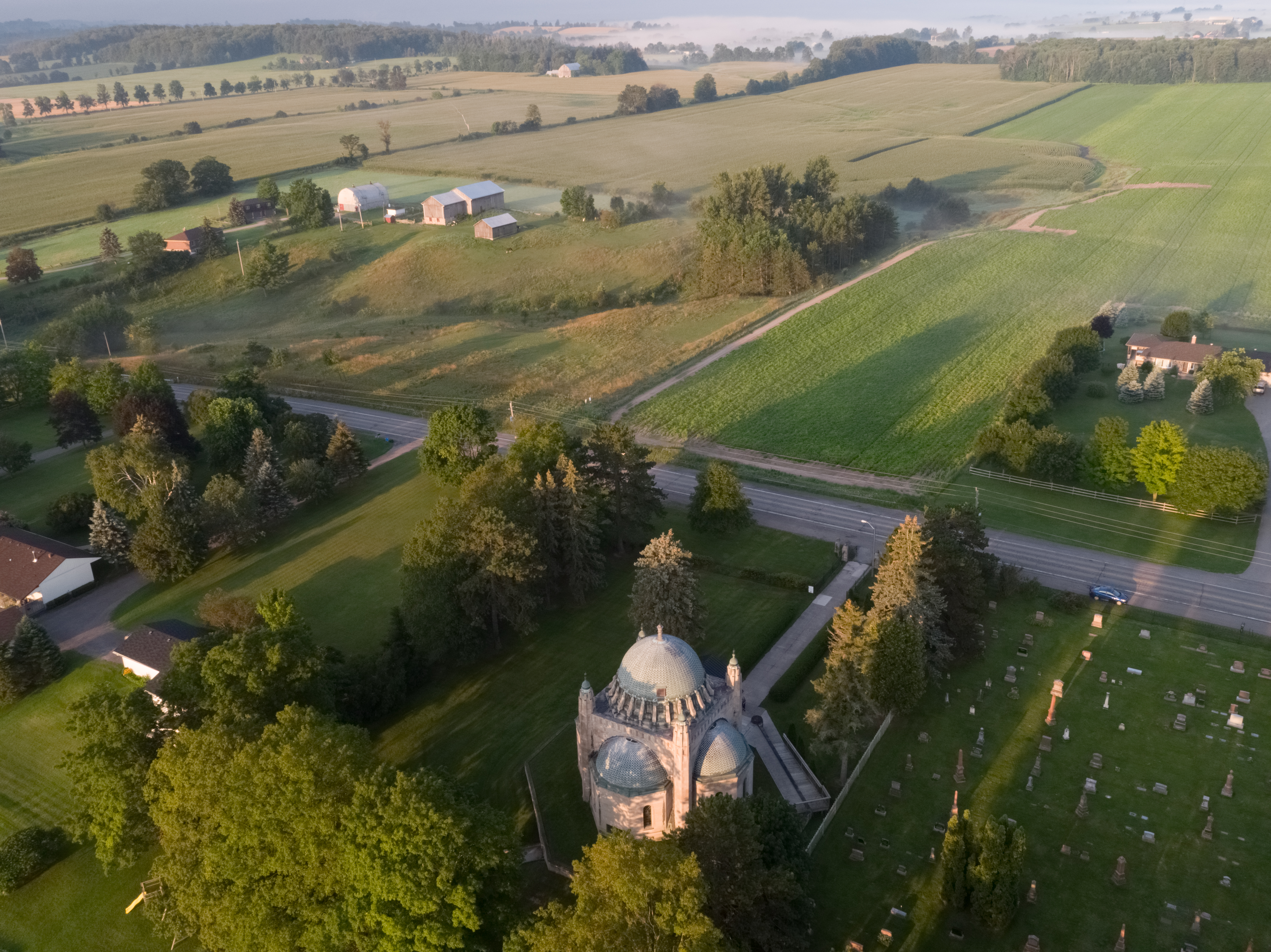 Aerial photo of the Thomas Foster Memorial in Uxbridge on a mostly sunny day with fog lifting in the background. The Thomas Foster is surrounded by farms and trees in a rural setting.