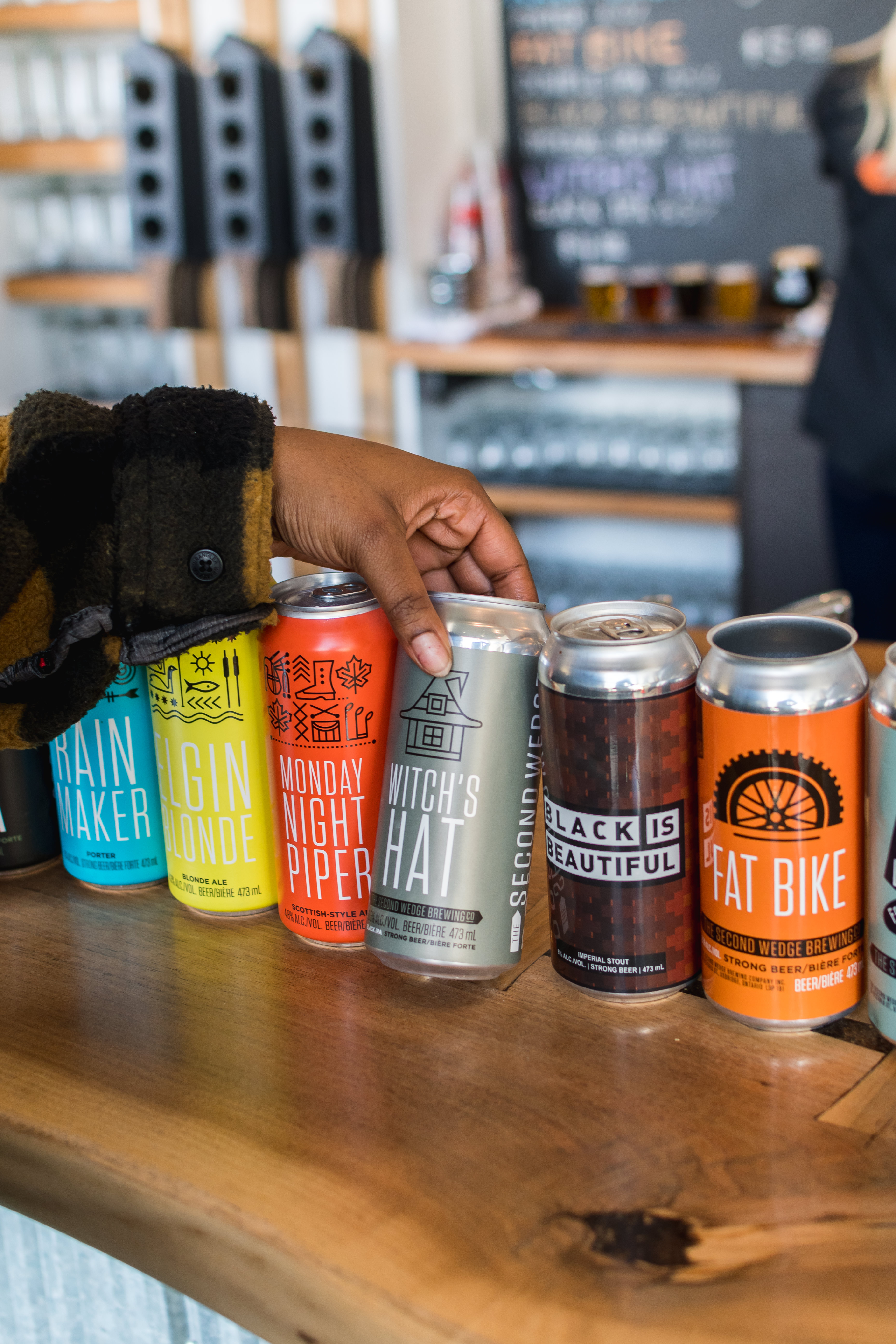 Image includes an arm picking up a grey can of Witch's Brew beer made by the Second Wedge Brewing Company. The can is sitting on a bar counter next to a blue can of Elgin Blonde, a yellow can of Rain Maker, an orange can of Monday Night Piper, a black can of Black is Beautiful and an orange can of Fat Tire. All of these are beers made by the Second Wedge Brewing Company.