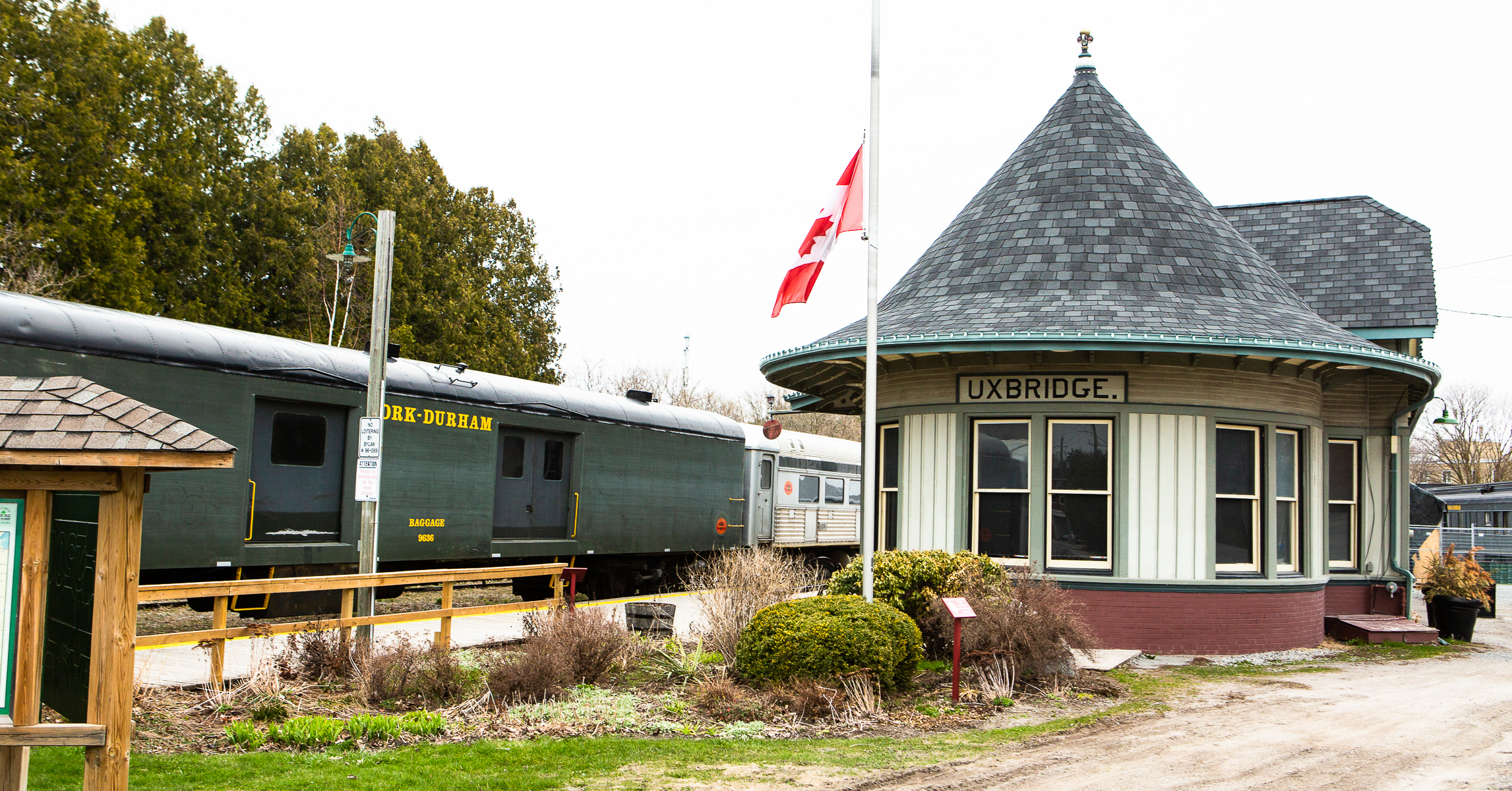 Image is of the York Durham Heritage Railway train station that has a roof that looks like a witch hat. There is a train with a baggage car parked near the station. A Canadian flag waves on a pole and the background is lined with trees.