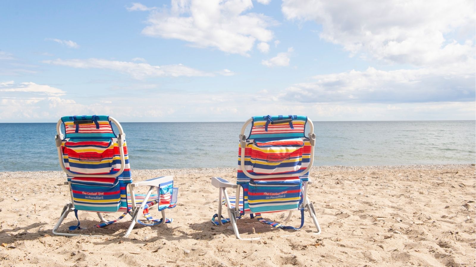 Image of two colourful striped lawn chairs facing the water on a beach