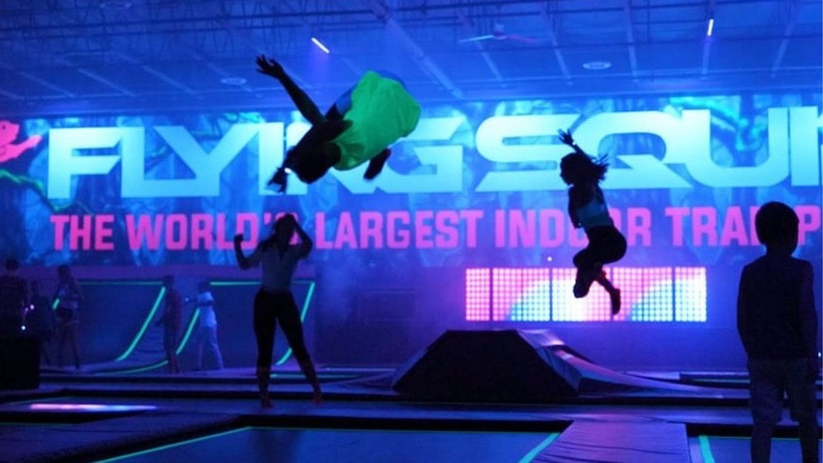 Image of an indoor trampoline park with black lights on and several people in all different stages of jumping