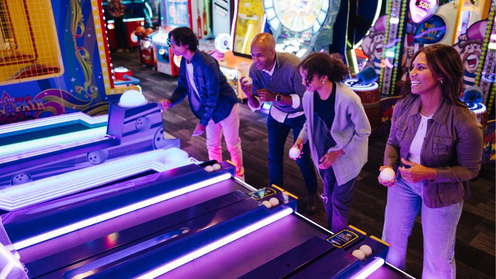Image of a family at an arcade playing skee-ball with brightly lit arcade games in the background