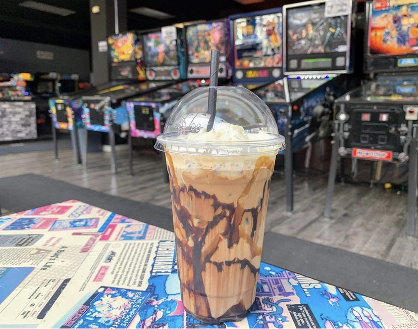 An iced coffee sits on a table with arcade games behind it