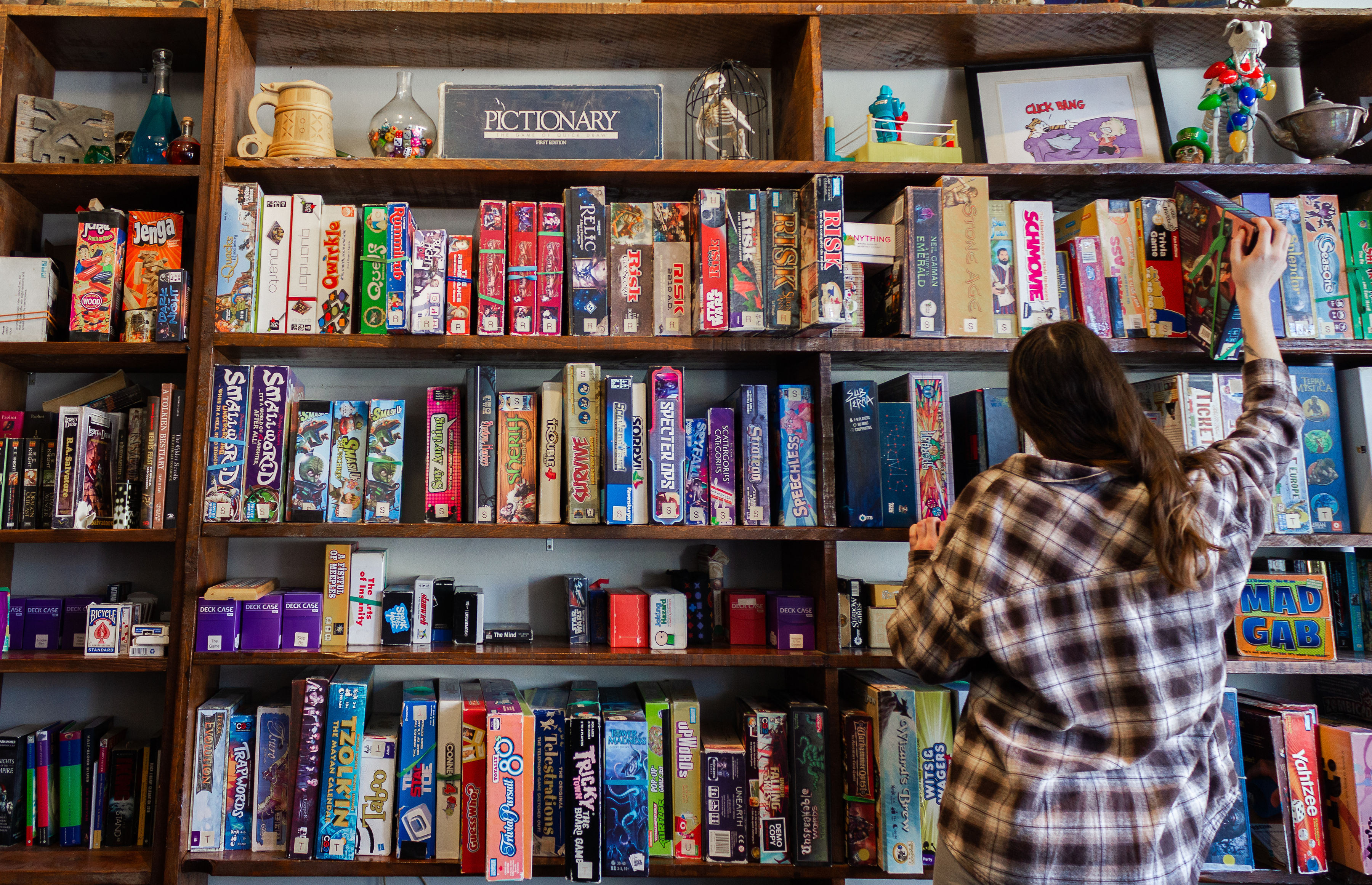 A person standing in front of a wall full of board games, reaching for one on the top shelf