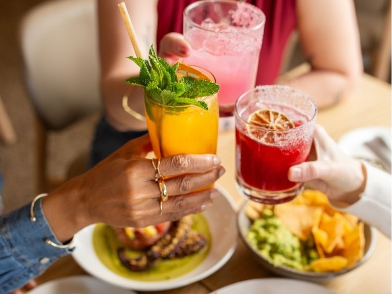 Image of three people clinking cocktail glasses over a dining table at a restaurant full of Mexican dishes