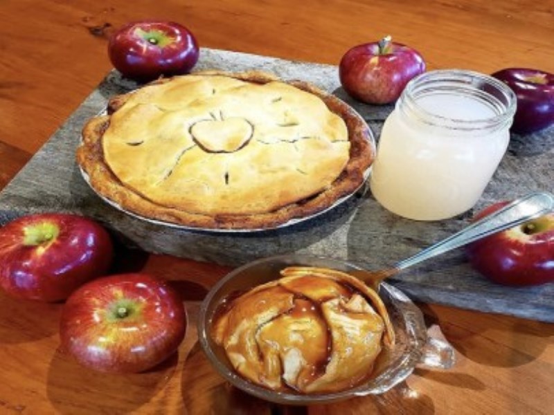 A baked apple pie surrounded by red apples and an apple dumping with caramel sauce