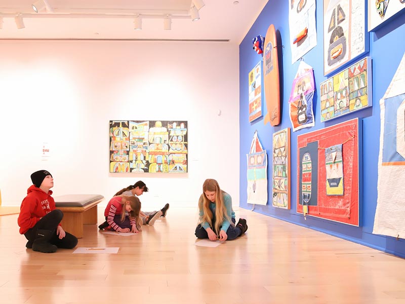 People sitting on the floor inside an art gallery admiring art displayed don the wall.
