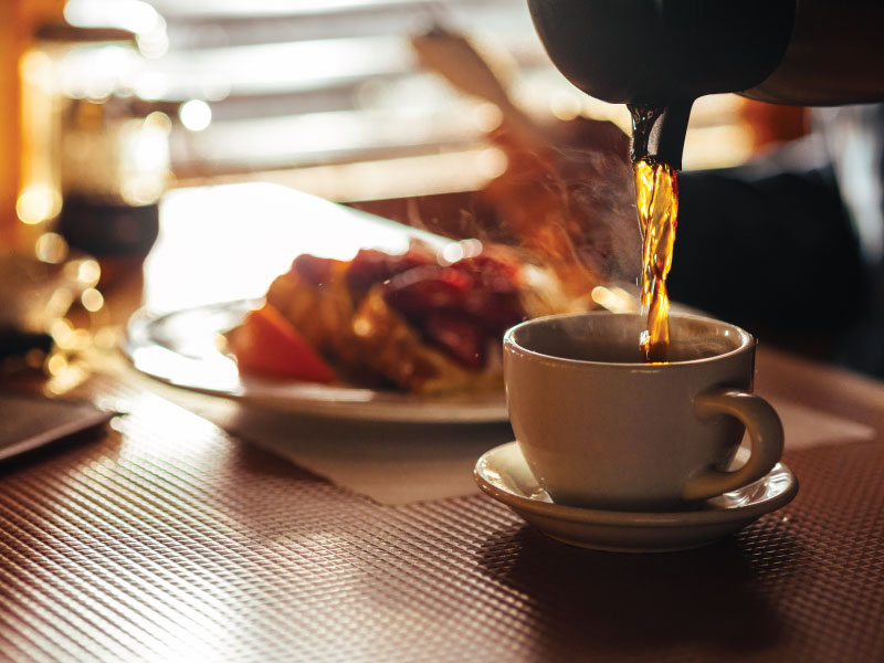 Close up of a cup of hot coffee on a table with a plate of breakfast in the background.