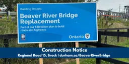 A sign announcing the Beaver River Bridge Replacement, with the temporary Beaver River Bridge in the background. The text below the image states, “Construction Notice, Regional Road 15, Brock, durham.ca/BeaverRiverBridge.”