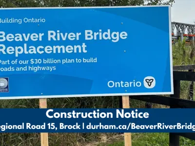 A sign announcing the Beaver River Bridge Replacement, with the temporary Beaver River Bridge in the background. The text below the image states, “Construction Notice, Regional Road 15, Brock, durham.ca/BeaverRiverBridge.”
