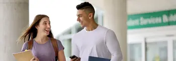 Two people are walking and talking while holding folders and a phone. They appear to be near a Student Services building.
