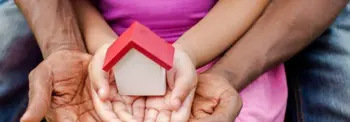 Two pairs of hands gently hold a small model house with a red roof. The house rests in the larger hands, with a pair of smaller hands on top, against a blurred outdoor background.