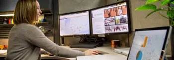 A person sits at a desk, interacting with two computer monitors displaying a map and a website about Durham. Another laptop is open with a digital map shown. A plant and office supplies are visible nearby.