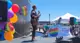 A person on a stage speaking into a microphone with Pride Balloons in the background.