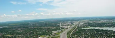 Aerial view of a large suburban area with a multi-lane highway running through it, surrounded by dense neighborhoods, green spaces, and a small lake under a partly cloudy blue sky.