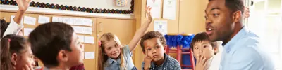 A group of young children sit on the floor in a classroom, attentively listening to an educator.