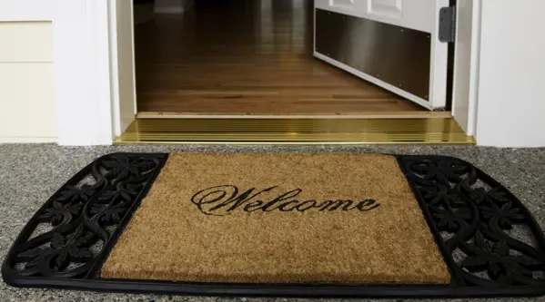 Close-up of a front doorway with an open door, showing a “Welcome” doormat on a porch in front of a wooden floor interior.