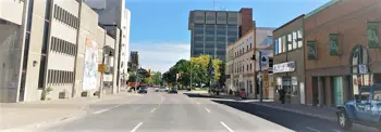 A view of a street featuring buildings on both sides. On the left, there's a large concrete structure with murals, and on the right, a series of brick buildings, including businesses and a restaurant. In the background, a tall office building is visible. The street is lined with trees, and a few vehicles are on the road. The sky is clear and blue.