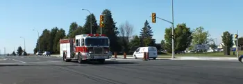 A fire truck and a white van are stopped at a red traffic light at an intersection. There are traffic lights, orange cones visible, and trees school in the background.
