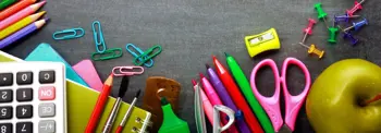 Brightly coloured school supplies arranged on a dark surface, including pencils, pens, scissors, paper clips, push pins, a calculator, a sharpener, notebooks, a ruler, brushes, and an apple.
