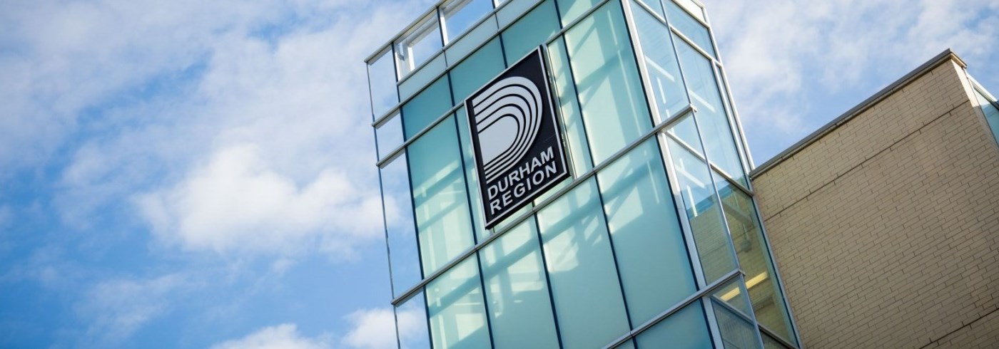 A modern glass building facade with a prominent sign displaying the Durham Region logo in white, on a dark navy background. 